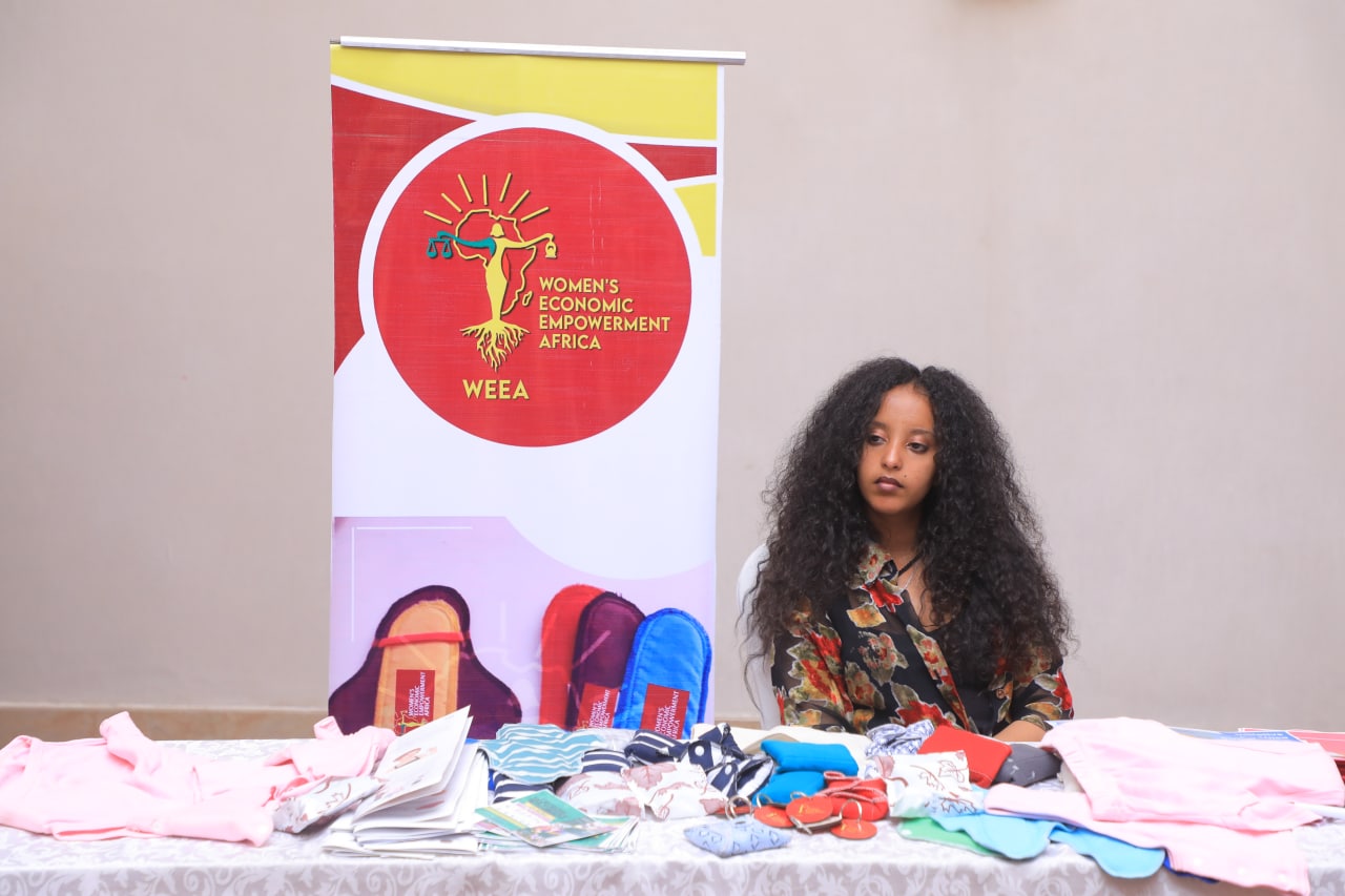 Reusable menstrual products and learning materials displayed on a table beside a WEEA banner.
