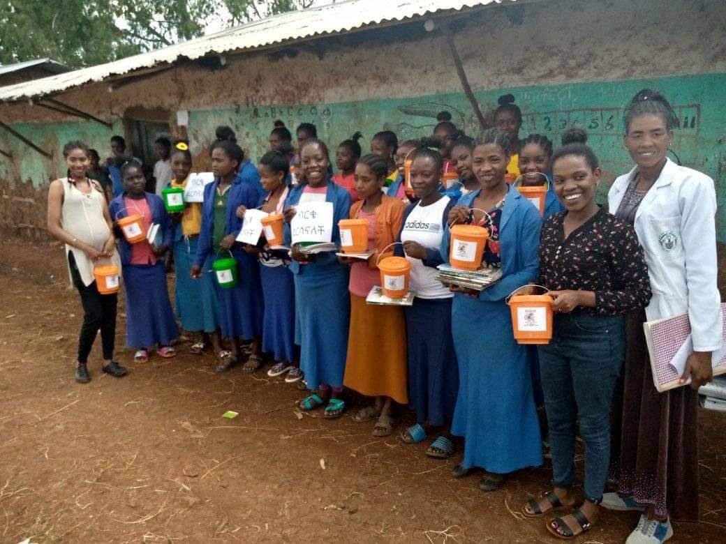 Participants holding dignity buckets and learning materials outside a school building.