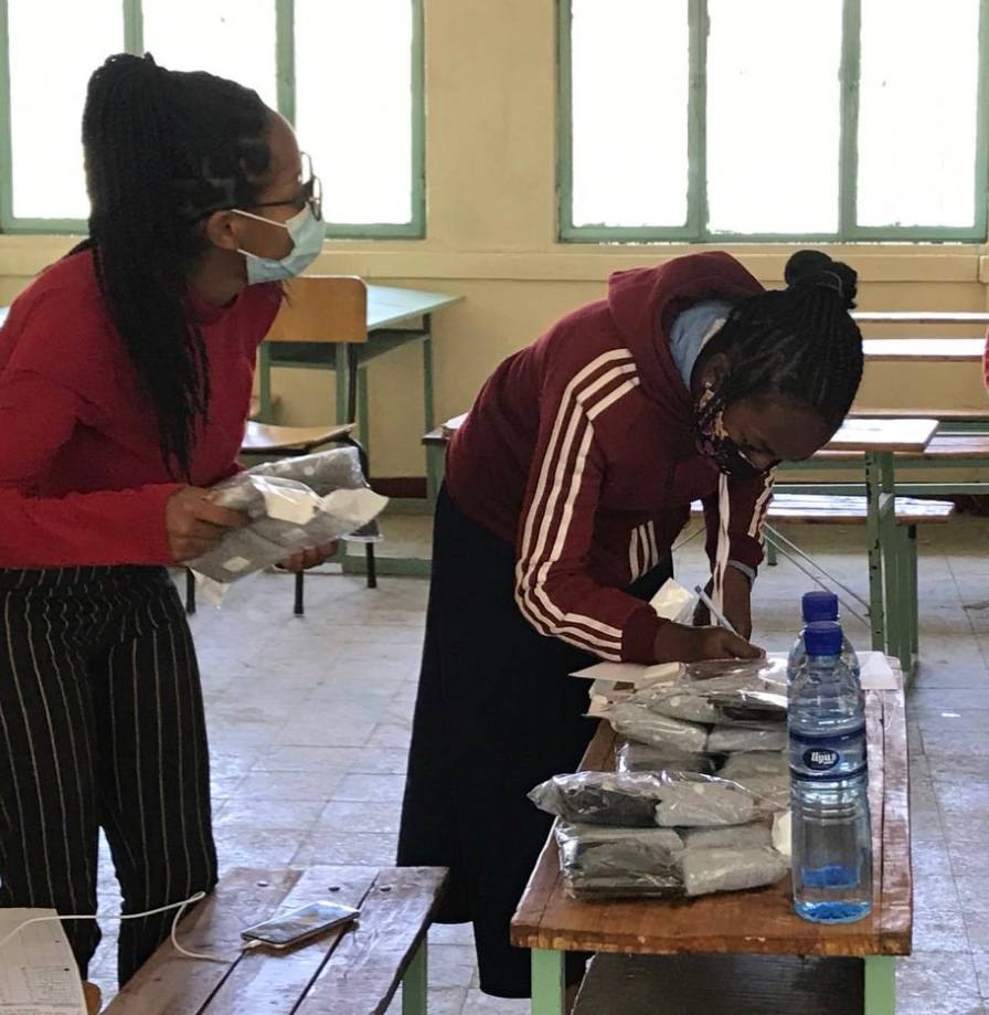 Two people preparing packaged supplies beside a classroom desk.