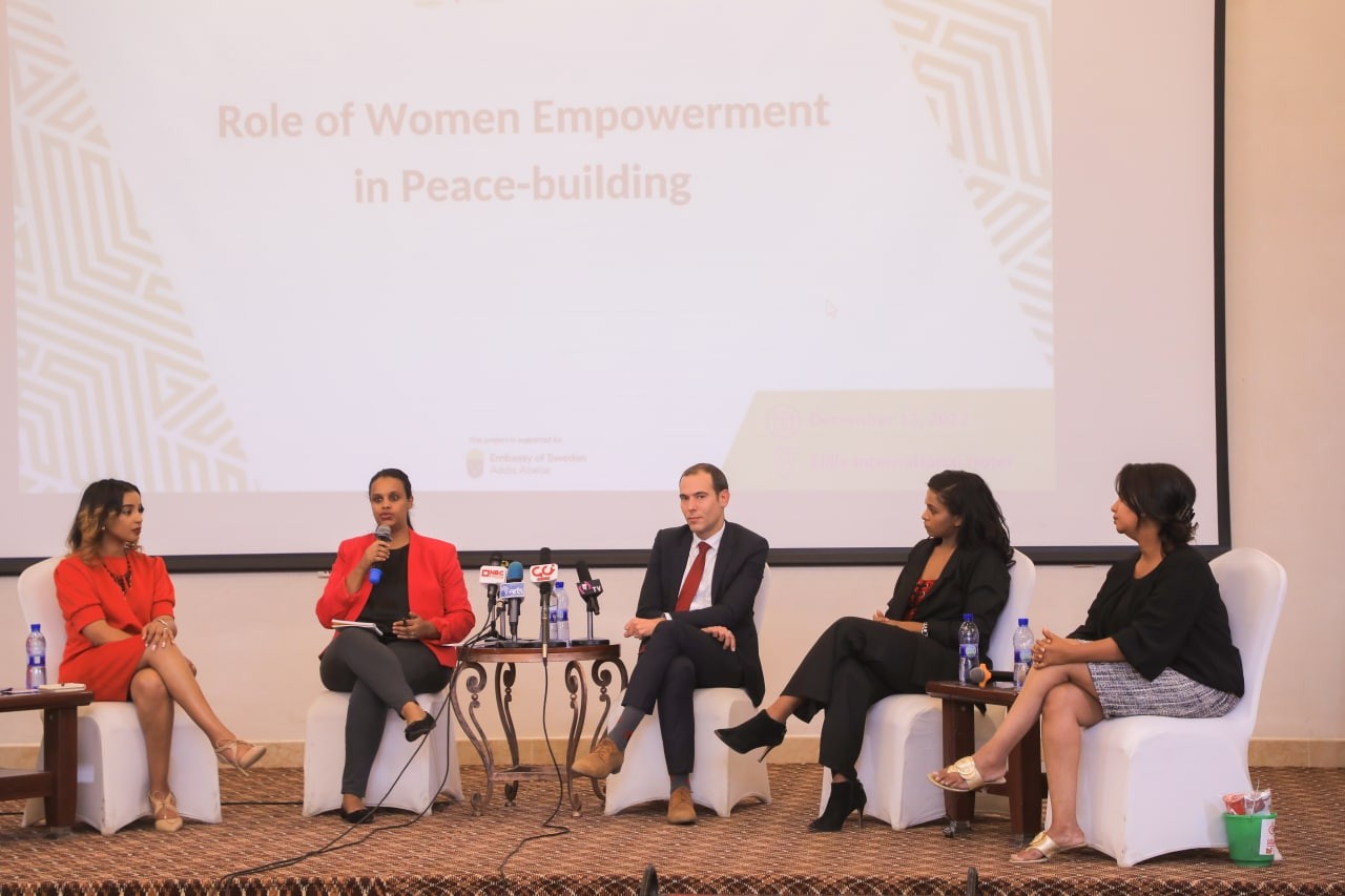 Panelists seated under a slide about women’s empowerment and peacebuilding, with one speaking into a microphone.